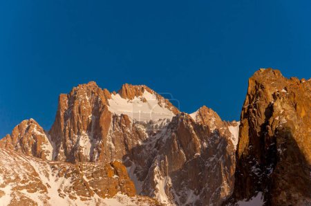 Téléchargez les photos : Les falaises brun rougeâtre du pic Korona s'élèvent précipitamment, leurs crêtes enneigées gravées contre le ciel bleu profond, un défi réputé pour les grimpeurs et un spectacle pour les touristes en visite - en image libre de droit