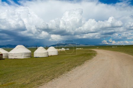Téléchargez les photos : Une rangée de yourtes kirghizes traditionnelles parsèment les prairies ouvertes de Son-Kul, avec des montagnes lointaines encadrant l'horizon, sous un vaste ciel marqué par des traînées de nuages blancs, reflétant un patrimoine nomade. - en image libre de droit