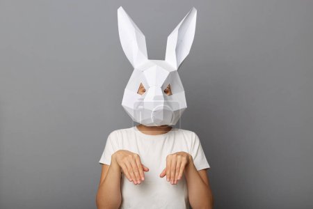 Téléchargez les photos : Portrait d'une drôle de femme inconnue portant un masque de lapin et un t-shirt blanc debout avec des pattes de lièvre, exprimant des émotions festives, isolée sur fond gris. - en image libre de droit