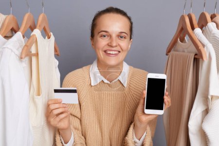 Photo for Indoor shot of smiling cheerful woman standing near clothes hang on shelf, showing mobile phone with empty display and credit card, presenting copy space for advertisement area. - Royalty Free Image