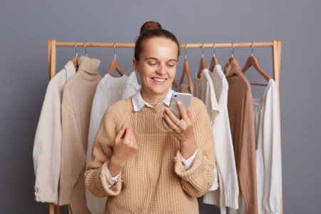 Photo for Image of pleased Caucasian young adult woman using cell phone, reading messages, smiling happily, wearing beige sweater, standing near clothes hang on shelf. - Royalty Free Image