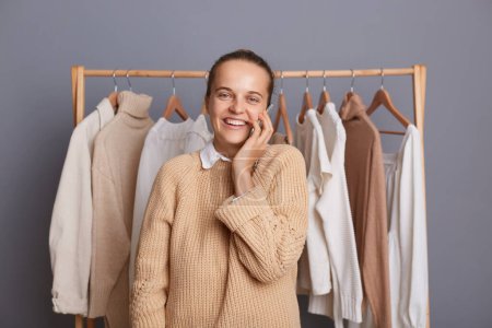 Photo for Indoor shot of attractive joyful happy Caucasian woman standing near clothes hang on shelf and talking on smart phone with her new client or buyer, looking at camera. - Royalty Free Image