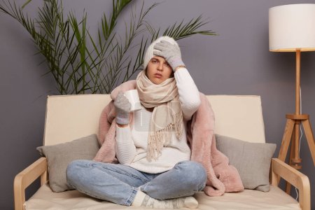 Traurige ungesunde kranke Frau, die zu Hause mit Wintermantel, Hut und Handschuhen auf dem Sofa sitzt, sich unwohl fühlt, im kalten Haus posiert, Kopfschmerzen hat, heißen Tee trinkt, Grippesymptome hat.