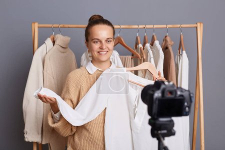 Téléchargez les photos : Portrait de heureuse femme souriante enchantée debout près de cintre rack avec des vêtements dans le magasin de mode ou à la maison dans la garde-robe, montrant chemisier blanc à ses disciples, la vente de tenues. - en image libre de droit