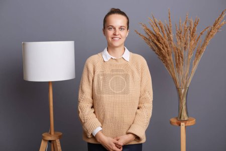 Photo for Indoor shot of pretty positive woman wearing beige sweater standing against gray wall with lamp and dried flowers, looking at camera with toothy smile, being in good mood. - Royalty Free Image