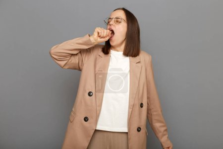 Photo for Drowsy tired young adult woman with dark hair wearing jacket and glasses posing against gray wall, standing with wide open mouth, yawning feeling fatigued, lack of energy, need sleep and rest. - Royalty Free Image