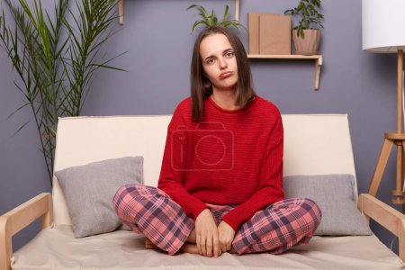 Photo for Indoor shot of tired exhausted sad bored brown-haired woman wearing red sweater and checkered pants sitting on cough in living room at home, looking at camera, needs entertainment. - Royalty Free Image