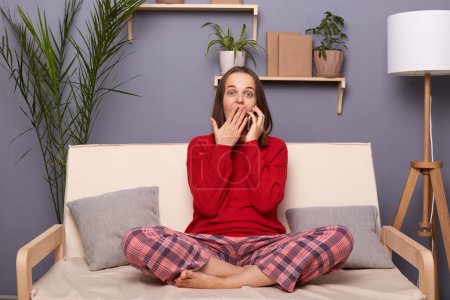 Photo for Indoor shot of shocked astonished brown-haired female wearing red sweater and checkered pants sitting on cough in room at home, talking on smart phone, hearing breaking news, covering mouth with palm. - Royalty Free Image