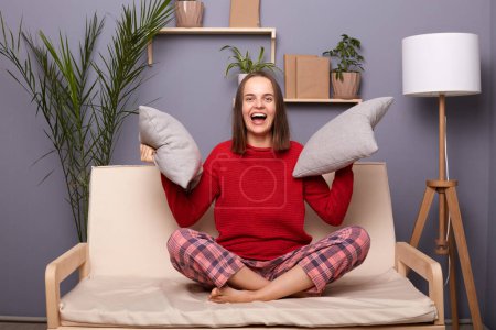 Photo for Photo of positive joyful cheerful Caucasian female dressed in home clothes holding two pillows in hands, looking at camera with toothy smile, resting with good mood, sitting on cough in her flat. - Royalty Free Image