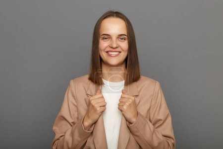 Photo for Indoor shot of beautiful smiling joyful young adult woman wearing beige jacket standing with toothy smile, enjoying, being in good mood, posing isolated on gray background. - Royalty Free Image
