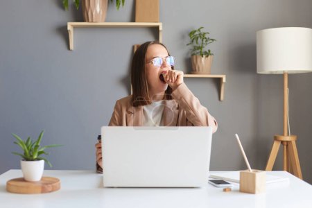 Photo for Attractive beautiful young Caucasian female feel tired and overworked, woman in glasses work remotely from home or sitting at table in office in front of laptop, yawning and covering mouth. - Royalty Free Image