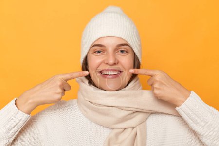 Photo for Happy woman wearing white sweater, cap and scarf, pointing index fingers at toothy smile, showing well cared white teeth after whitening or dentist visit, standing isolated over yellow background. - Royalty Free Image