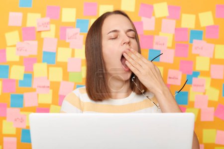 Photo for Tired sleepy bored young adult woman with brown hair working on laptop, keeps eyes closed, covering mouth and yawning, working on computer long hours, posing against yellow wall with sticky notes. - Royalty Free Image