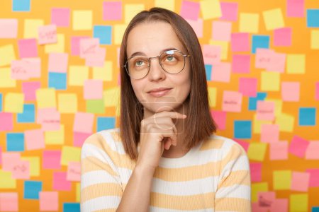 Photo for Indoor shot of pensive thoughtful woman with brown hair in striped t shirt standing against yellow wall with colorful stickers, holding her chin, looking away, dreaming. - Royalty Free Image