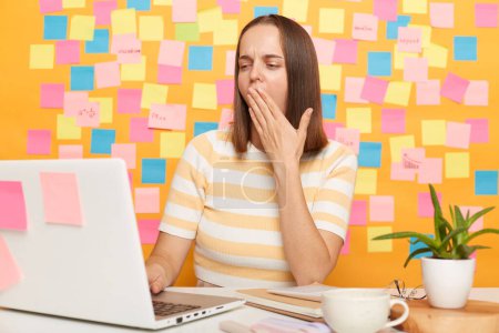 Photo for Indoor shot of sleepy tired exhausted woman sitting at table, working on laptop against yellow wall covered with sticky notes, covering mouth with palm, being sleepless. - Royalty Free Image