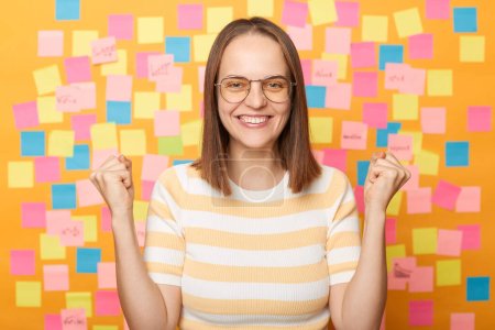 Photo for Happy excited woman with brown hair standing isolated over yellow background with stickers, celebrating her winning, clenched fists, looking at cameras with toothy smile. - Royalty Free Image