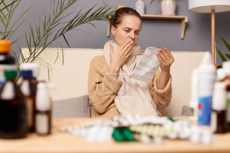 Photo for Indoor shot of young adult woman with medicine and pills, ill female looking at medication explanation before taking prescription drugs, wearing sweater and scarf, covering mouth with palm. - Royalty Free Image