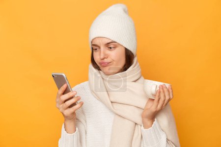 Photo for Image of sad upset attractive young adult woman wearing warm sweater, cap and scarf posing, holding cup of coffee, using cell phone, checking social networks, isolated over yellow background. - Royalty Free Image