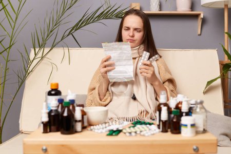Photo for Indoor shot of confused sick unhealthy woman reading instructions before taking medication while sitting on sofa at home, catching cold, shaving flu symptoms. - Royalty Free Image