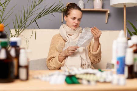 Photo for Indoor shot of sick young adult woman sitting on sofa wrapped in scarf surrounded with medicines, posing in living room at home, reading instruction before taking medicine. - Royalty Free Image