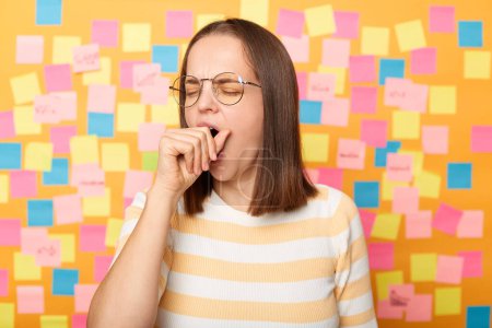 Photo for Portrait of sleepy overworked attractive woman wearing striped T-shirt standing against yellow wall covered with stickers, yawning, covering mouth with fist, keeps eyes closed. - Royalty Free Image