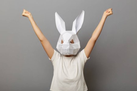 Téléchargez les photos : Portrait d'une femme ravie portant un t-shirt blanc et un masque de lapin en papier debout avec les bras levés, célébrant sa victoire, posant isolée sur fond gris. - en image libre de droit