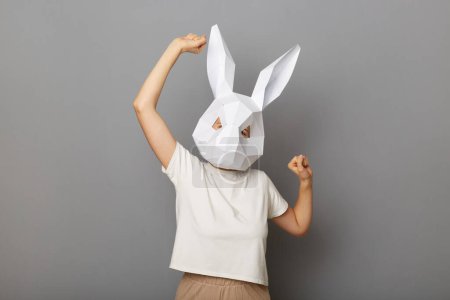 Téléchargez les photos : Portrait d'une femme festive extrêmement heureuse portant un t-shirt blanc et un masque de lapin en papier, posant les bras levés, dansant, étant de bonne humeur, se tenant isolée sur un fond gris. - en image libre de droit