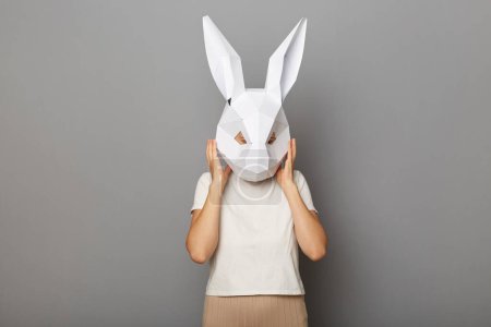 Horizontal shot of woman wearing white t shirt and paper rabbit mask standing isolated over gray background, taking off her mask or covering ears, mind blowing.