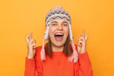 Photo for Image of excited happy woman wearing orange sweater and knitted earflap hat standing isolated over yellow background, dreaming about future, crossing her fingers. - Royalty Free Image