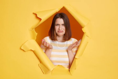Photo for Portrait of despair young woman wearing striped T-shirt breaking through hole in yellow paper, being offended and upset, raised arms, doesn't know how it happened. - Royalty Free Image