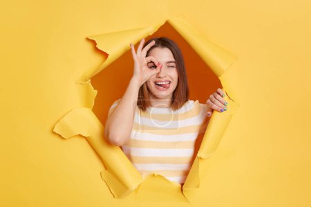 Photo for Horizontal shot of satisfied playful beautiful woman wearing striped T-shirt looking through hole in yellow paper wall, showing ok gesture, covering eye with okay sign. - Royalty Free Image