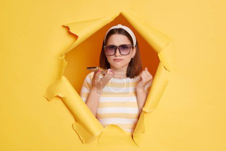 Photo for Portrait of angry woman wearing striped T-shirt, hair band and sunglasses, holding mobile phone, recording voice message, keeps teeth clenched, breaking through paper hole in yellow wall. - Royalty Free Image