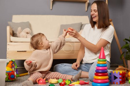 Foto de Indoor shot of mom and daughter together sitting on floor near toough and playing with toys, mommy having fun with her toddler kid, expressed positive emotions. - Imagen libre de derechos