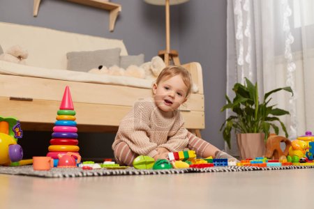 Téléchargez les photos : Portrait de mignon enfant féminin souriant portant pull beige assis sur le sol entouré de jouets colorés, regardant la caméra, jouant à la maison, souriant joyeusement. - en image libre de droit