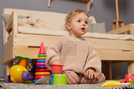 Téléchargez les photos : Plan intérieur de mignonne petite fille joyeuse portant un pull beige assis sur le sol près du canapé entouré de jouets en plastique colorés, regardant loin avec la bouche ouverte, voit quelque chose d'intéressant. - en image libre de droit