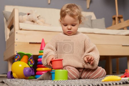 Téléchargez les photos : Portrait de petite charmante petite fille portant un pull beige jouant avec des jouets en plastique colorés, assise sur le sol, s'amusant et. Apprentissage précoce. Jouets développement. - en image libre de droit