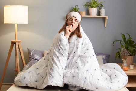 Photo for Indoor shot of sleepy bored tired woman in sleeping mask sitting on bed wrapped in duvet, yawning covering mouth with hand, needs to have nap, being sleepless due lack of sleep. - Royalty Free Image