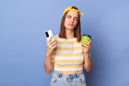 Photo for Portrait of sad sleepy tired woman wearing casual style clothing standing isolated over blue background, holding takeaway coffee and mobile phone, being unhappy. - Royalty Free Image