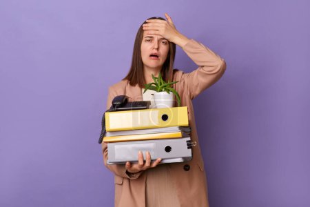 Photo for Horizontal shot of sad crying woman wearing beige jacket holding lot of documents folders isolated over lilac background., showing facepalm gesture, being fired. - Royalty Free Image