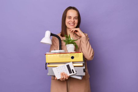 Photo for Indoor shot of smiling beautiful woman wearing beige jacket holding lot of documents folders, looking at camera with toohy smile, keeps hand under chin, posing isolated over purple background. - Royalty Free Image