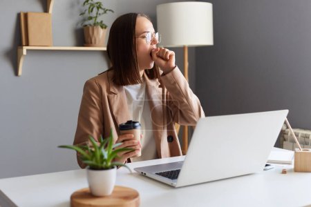 Foto de Portrait of tire exhausted sleepy woman with brown hair wearing beige jacket working online on laptop, yawning, covering mouth with palms, drinking coffee. - Imagen libre de derechos