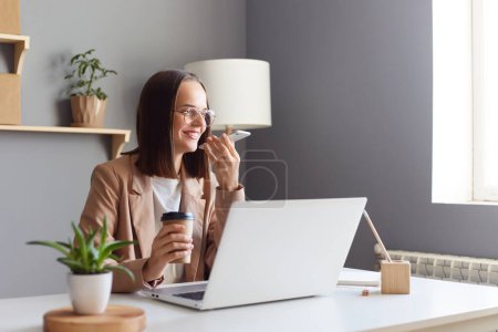 Photo for Horizontal shot of joyful cheerful woman with brown hair wearing beige jacket working online on laptop, drinking coffee and recording voice message, expressing positive emotions. - Royalty Free Image