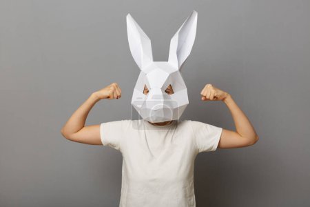 Téléchargez les photos : Image of powerful woman wearing white T-shirt and paper rabbit mask standing isolated over gray background, raised her arms, demonstrates her power, showing how strong she is. - en image libre de droit
