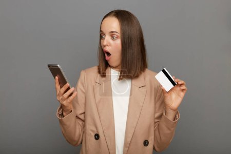 Photo for Indoor shot of amazed astonished woman with brown hair wearing beige jacket posing isolated over gray background, holding credit card in hand and using cell phone, being very surprised. - Royalty Free Image