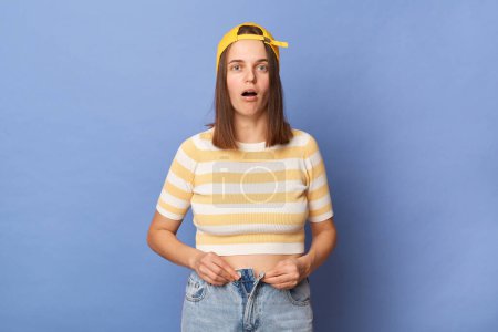 Photo for Portrait of shocked sad teen girl wearing striped T-shirt and baseball cap standing isolated over blue background, female looking at camera, need to lose weight. - Royalty Free Image