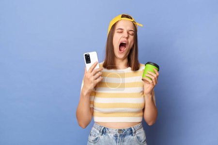 Photo for Horizontal shot of sleepy tired teen girl wearing striped T-shirt and baseball cap standing isolated over blue background, holding coffee to go and cell phone, yawning, needs energy. - Royalty Free Image