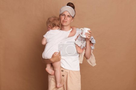 Téléchargez les photos : Image of sad depressed woman wearing white T- shirt holding her baby daughter in hands isolated over brown background, being exhausted to be with kid every day,feels fatigue during maternity leave. - en image libre de droit