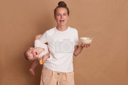 Téléchargez les photos : Portrait of shocked crying screaming woman wearing white T- shirt standing with her little daughter in hands isolated over brown background, trying to feed her child. - en image libre de droit