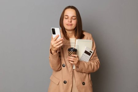 Photo for Photo of confused Caucasian woman wearing beige jacket holding organizer and calculator isolated over gray background, looking at mobile phone display with puzzled sleepy expression. - Royalty Free Image
