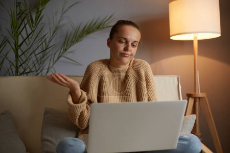 Téléchargez les photos : Indoor shot of puzzled confused Caucasian woman wearing beige jumper and jeans sitting on cough in lotus pose, having video call on laptop, shrugging shoulders, looks helpless. - en image libre de droit